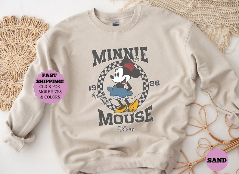 May include: Beige sweatshirt featuring a distressed graphic of Minnie Mouse. The design includes the text "MINNIE MOUSE" and the year "1928". The image also includes the text "CLICK FOR MORE SIZES & COLORS".