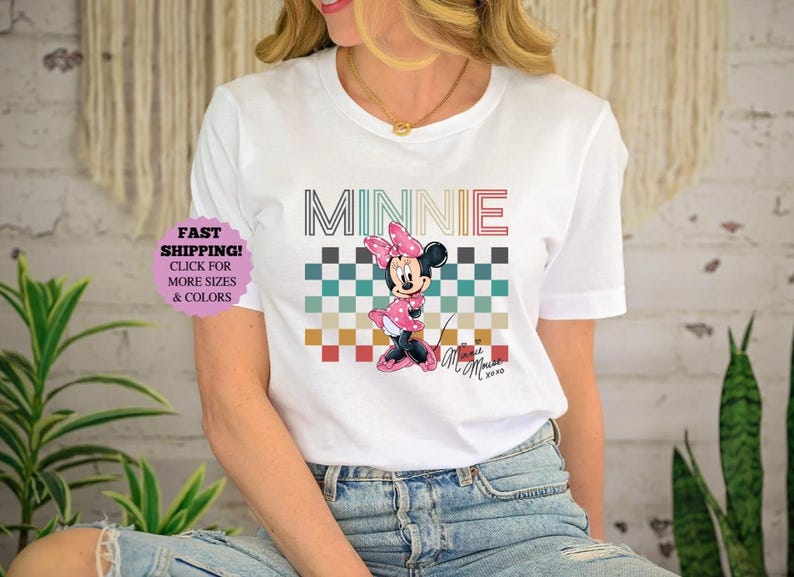以下が含まれることがあります： ミニーマウスのグラフィックが特徴の白いTシャツ。デザインには、マルチカラーのフォントで書かれた「MINNIE」の文字、市松模様、ピンクのドレスを着たミニーマウスの漫画の画像が含まれています。