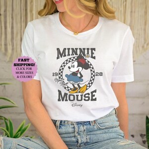 May include: White t-shirt featuring a vintage-style Minnie Mouse graphic. The design includes Minnie Mouse in a blue skirt and yellow shoes, with the text "MINNIE MOUSE" and the year "1928". The Disney logo is at the bottom.