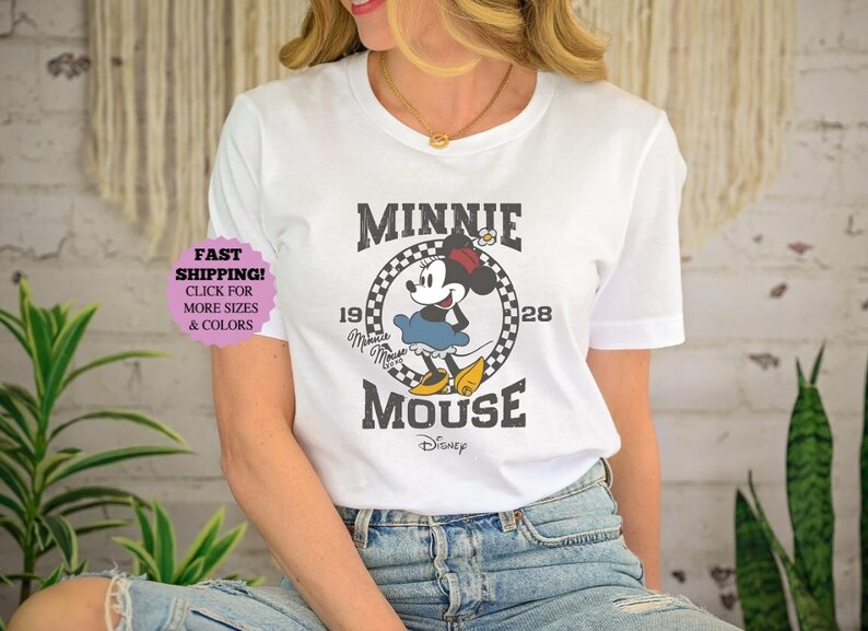 May include: White t-shirt featuring a vintage-style Minnie Mouse graphic. The design includes Minnie Mouse in a blue skirt and yellow shoes, with the text "MINNIE MOUSE" and the year "1928". The Disney logo is at the bottom.