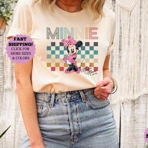 以下が含まれることがあります： ミニーマウスのグラフィックが特徴のクリーム色のTシャツ。デザインには、レトロなフォントで書かれた「MINNIE」の文字、市松模様、ミニーマウスのイラストが含まれています。このシャツは半袖で、デニムジーンズと組み合わせています。