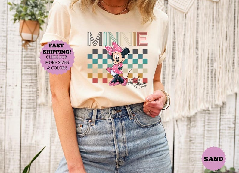 以下が含まれることがあります： ミニーマウスのグラフィックが特徴のクリーム色のTシャツ。デザインには、レトロなフォントで書かれた「MINNIE」の文字、市松模様、ミニーマウスのイラストが含まれています。このシャツは半袖で、デニムジーンズと組み合わせています。