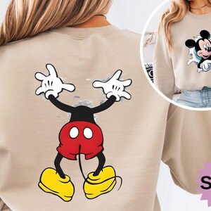 Puede incluir: Sudadera color arena con un gr&aacute;fico de Mickey Mouse. La parte delantera muestra a Mickey rompiendo un efecto de papel rasgado, mientras que la parte trasera muestra una imagen de cuerpo completo de Mickey Mouse con los brazos levantados. La sudadera tiene cuello redondo.