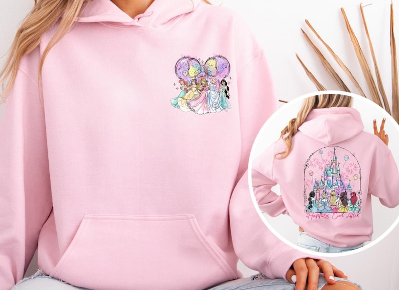 May include: A light pink hoodie with a front pocket and a drawstring hood. The front features a colorful graphic of cartoon princesses. The back of the hoodie has a castle graphic with the text "Happily Ever After".