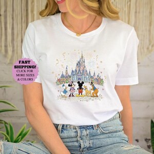 May include: White t-shirt featuring a colorful illustration of a castle and cartoon characters. The design includes Mickey Mouse, Minnie Mouse, Donald Duck, Goofy, and Pluto. The shirt has the text "FAST SHIPPING! CLICK FOR MORE SIZES & COLORS".