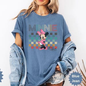 May include: Blue jean-colored t-shirt featuring a graphic of Minnie Mouse. The shirt has the word "MINNIE" across the top in a multi-colored font. Minnie Mouse is wearing a pink polka-dot dress and bow. The shirt is a Comfort Color brand.
