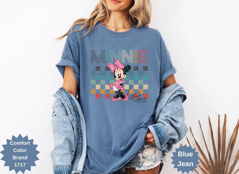 May include: Blue jean-colored t-shirt featuring a graphic of Minnie Mouse. The shirt has the word "MINNIE" across the top in a multi-colored font. Minnie Mouse is wearing a pink polka-dot dress and bow. The shirt is a Comfort Color brand.