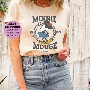 May include: Cream-colored t-shirt featuring a vintage-style Minnie Mouse graphic. The design includes Minnie Mouse in a blue dress, yellow shoes, and a red bow, with the text "MINNIE MOUSE" and "1928".