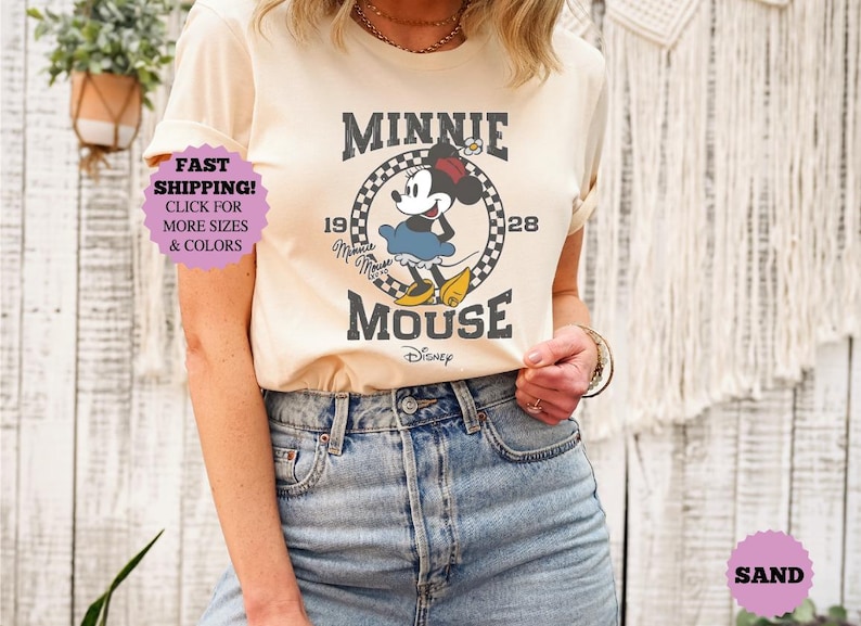 May include: Cream-colored t-shirt featuring a vintage-style Minnie Mouse graphic. The design includes Minnie Mouse in a blue dress, yellow shoes, and a red bow, with the text "MINNIE MOUSE" and "1928".