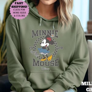 May include: Military green hoodie featuring a graphic of Minnie Mouse. The design includes the text "MINNIE MOUSE" and the years "19" and "28". The hoodie has a front pocket and a drawstring hood. The image also includes a pink sticker with the text "FAST SHIPPING! CLICK FOR MORE SIZES & COLORS".