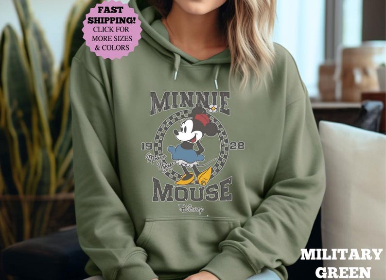 May include: Military green hoodie featuring a graphic of Minnie Mouse. The design includes the text "MINNIE MOUSE" and the years "19" and "28". The hoodie has a front pocket and a drawstring hood. The image also includes a pink sticker with the text "FAST SHIPPING! CLICK FOR MORE SIZES & COLORS".