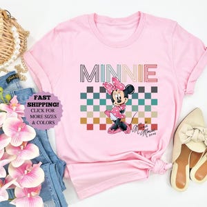 以下が含まれることがあります： ミニーマウスのグラフィックが特徴のピンクのTシャツ。デザインには、市松模様の上に「MINNIE」という名前が描かれています。ミニーマウスはピンクのドレスとリボンを着ています。画像にはジーンズと靴が含まれています。