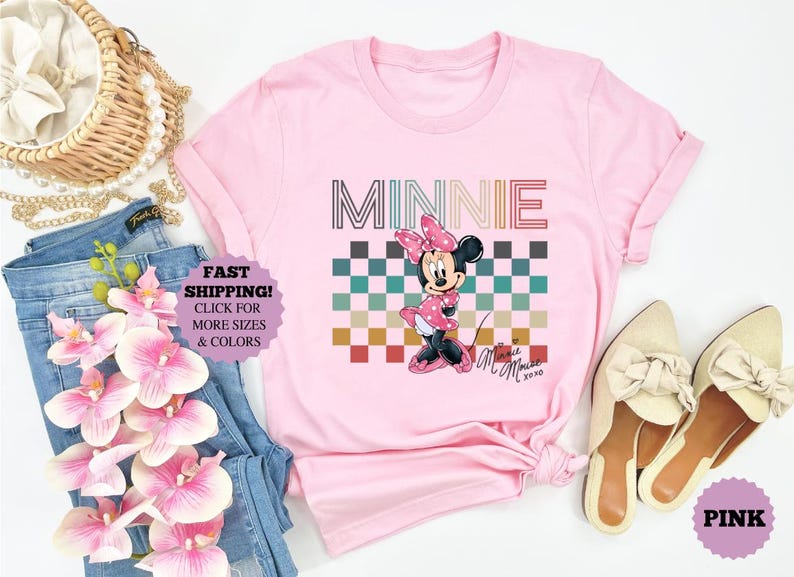 以下が含まれることがあります： ミニーマウスのグラフィックが特徴のピンクのTシャツ。デザインには、市松模様の上に「MINNIE」という名前が描かれています。ミニーマウスはピンクのドレスとリボンを着ています。画像にはジーンズと靴が含まれています。