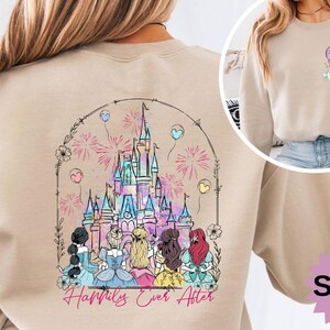 May include: Sand-colored sweatshirt featuring a colorful graphic of a fairytale castle with fireworks and balloons. The phrase "Happily Ever After" is written below the castle. A smaller graphic of princesses is on the upper right.