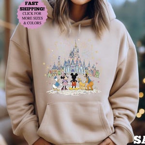 May include: Sand-colored hoodie featuring a colorful illustration of Disney characters in front of a castle. The design includes Mickey Mouse, Minnie Mouse, Donald Duck, Goofy, and a castle. The hoodie has a drawstring hood and a front pocket.