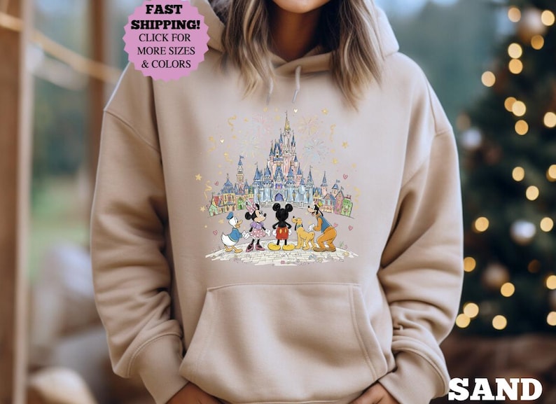 May include: Sand-colored hoodie featuring a colorful illustration of Disney characters in front of a castle. The design includes Mickey Mouse, Minnie Mouse, Donald Duck, Goofy, and a castle. The hoodie has a drawstring hood and a front pocket.