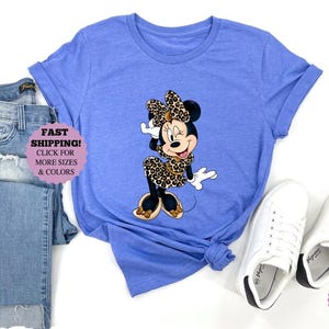May include: A heather blue t-shirt featuring a Minnie Mouse graphic in a leopard print dress and bow. The image includes a pair of blue jeans and white sneakers. The text "Fast Shipping!" is visible.