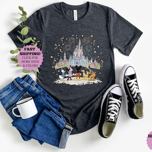 May include: Dark gray t-shirt featuring a colorful illustration of a castle, fireworks, and Disney characters. The shirt is paired with blue jeans, a white mug, and black canvas sneakers. The text "FAST SHIPPING!" is visible.