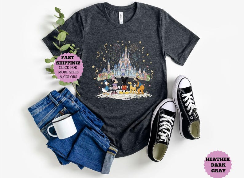 May include: Dark gray t-shirt featuring a colorful illustration of a castle, fireworks, and Disney characters. The shirt is paired with blue jeans, a white mug, and black canvas sneakers. The text "FAST SHIPPING!" is visible.