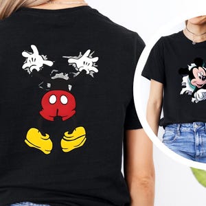 Puede incluir: Camiseta negra con un dise&ntilde;o de Mickey Mouse. La parte delantera muestra a Mickey Mouse saliendo de un agujero, y la parte trasera muestra sus manos, torso y pies. El dise&ntilde;o utiliza blanco, rojo, amarillo y negro.