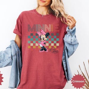 May include: Crimson-colored t-shirt featuring the word "MINNIE" in a retro font above a checkered pattern. The shirt displays a cartoon Minnie Mouse character in a pink dress and bow. The text "Minnie Mouse" is also visible.