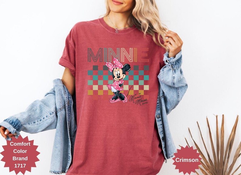 May include: Crimson-colored t-shirt featuring the word "MINNIE" in a retro font above a checkered pattern. The shirt displays a cartoon Minnie Mouse character in a pink dress and bow. The text "Minnie Mouse" is also visible.