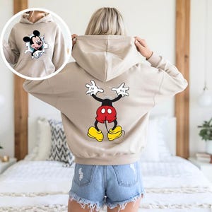 Puede incluir: Sudadera con capucha beige con gr&aacute;ficos de Mickey Mouse en la parte delantera y trasera. El gr&aacute;fico frontal muestra a Mickey rompiendo un agujero rasgado, mientras que la parte posterior muestra una imagen de cuerpo completo de Mickey Mouse con los brazos levantados. La sudadera tiene una capucha con cord&oacute;n.
