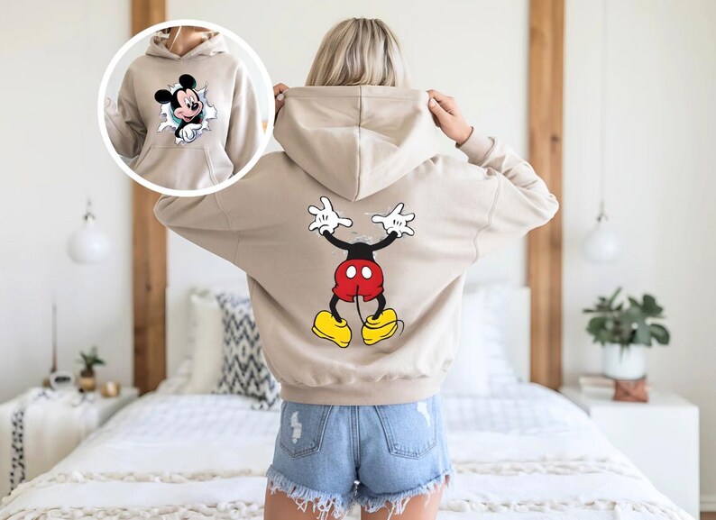 Puede incluir: Sudadera con capucha beige con gr&aacute;ficos de Mickey Mouse en la parte delantera y trasera. El gr&aacute;fico frontal muestra a Mickey rompiendo un agujero rasgado, mientras que la parte posterior muestra una imagen de cuerpo completo de Mickey Mouse con los brazos levantados. La sudadera tiene una capucha con cord&oacute;n.