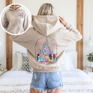 May include: Beige hoodie with a front pocket and a drawstring hood. The back features a colorful graphic of a castle, fireworks, and characters, with the text "Happily Ever After". A smaller graphic is on the front.