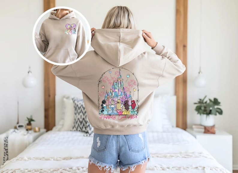 May include: Beige hoodie with a front pocket and a drawstring hood. The back features a colorful graphic of a castle, fireworks, and characters, with the text "Happily Ever After". A smaller graphic is on the front.