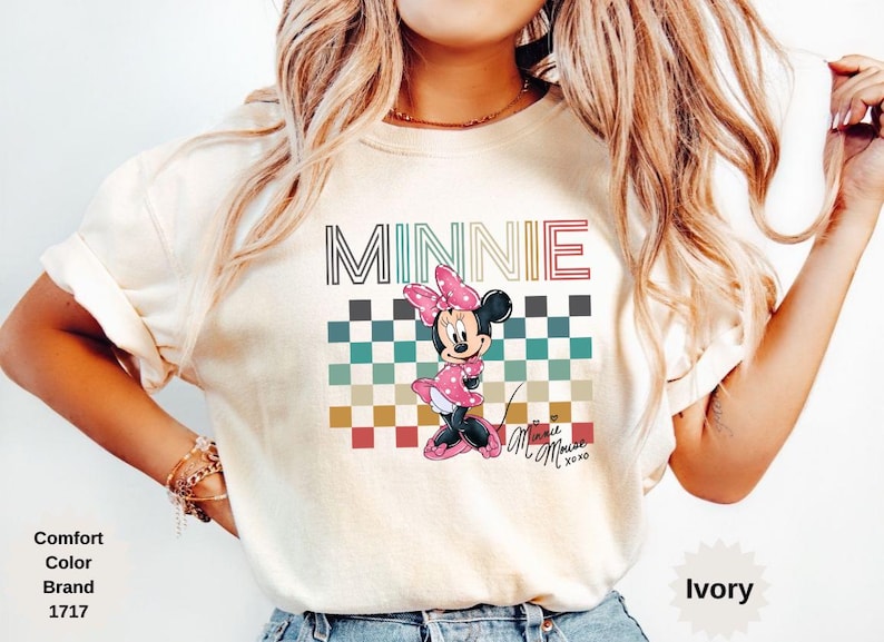 May include: Ivory-colored t-shirt featuring a Minnie Mouse graphic. The design includes the name "MINNIE" in a striped font above a checkered pattern and a cartoon Minnie Mouse wearing a pink dress and bow. The shirt has short sleeves.