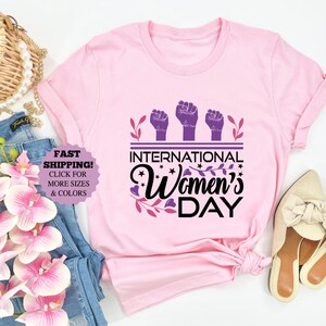 May include: A light pink t-shirt with the text "International Women's Day" in black and purple. The design includes three raised fists and floral accents. The shirt is displayed with jeans, shoes, and accessories.