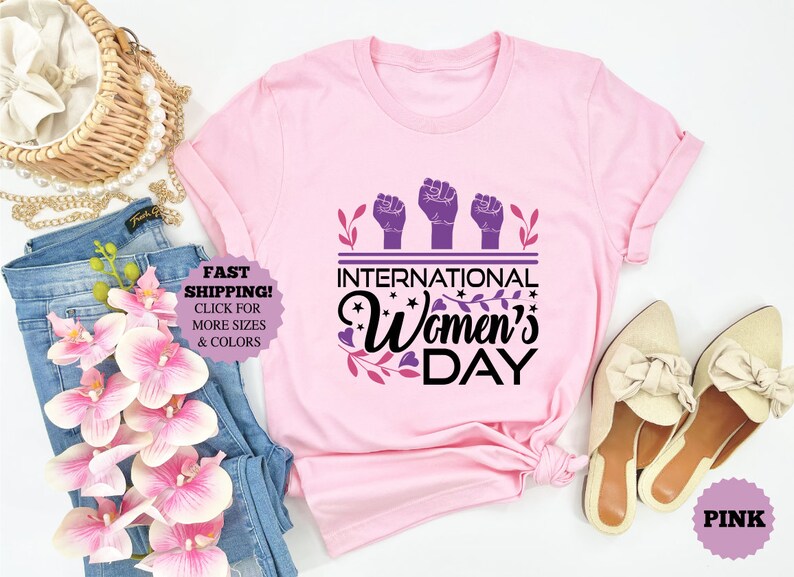 May include: A light pink t-shirt with the text "International Women's Day" in black and purple. The design includes three raised fists and floral accents. The shirt is displayed with jeans, shoes, and accessories.