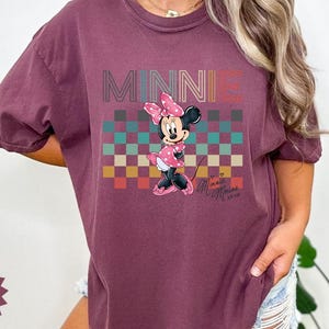 May include: Berry-colored t-shirt featuring a graphic of Minnie Mouse. The design includes the word "MINNIE" in a retro font above a checkered pattern. Minnie Mouse is depicted in a pink polka-dot dress and bow.