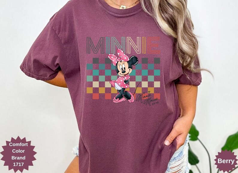 May include: Berry-colored t-shirt featuring a graphic of Minnie Mouse. The design includes the word "MINNIE" in a retro font above a checkered pattern. Minnie Mouse is depicted in a pink polka-dot dress and bow.