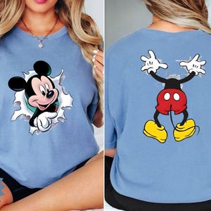 Peut inclure: T-shirt bleu clair avec Mickey Mouse. Le motif avant montre Mickey per&ccedil;ant un fond d&eacute;chir&eacute;, tandis que le dos affiche Mickey les bras lev&eacute;s. Le t-shirt porte les textes "Comfort Color Brand 1717" et "BlueJean".