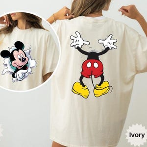 Peut inclure: T-shirt ivoire avec Mickey Mouse. L'avant pr&eacute;sente Mickey per&ccedil;ant un mur d&eacute;chir&eacute;, et le dos montre la pose embl&eacute;matique de Mickey. Le t-shirt porte le nom de la marque et le nom de la couleur.