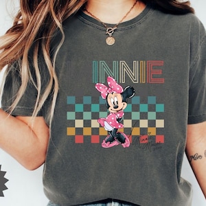 May include: Dark gray t-shirt featuring a graphic of Minnie Mouse with the word "INNIE" above her. Minnie is wearing a pink polka dot dress and bow. The background has a checkered pattern in various colors.