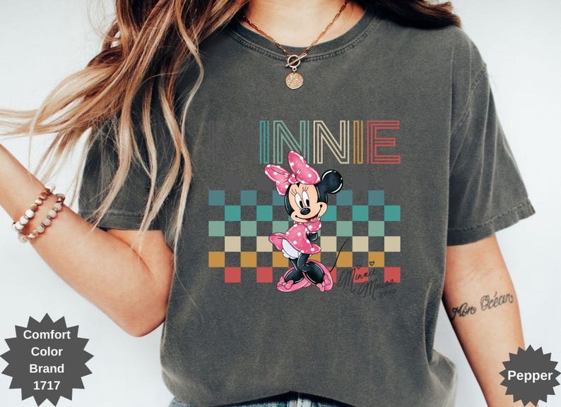 May include: Dark gray t-shirt featuring a graphic of Minnie Mouse with the word "INNIE" above her. Minnie is wearing a pink polka dot dress and bow. The background has a checkered pattern in various colors.