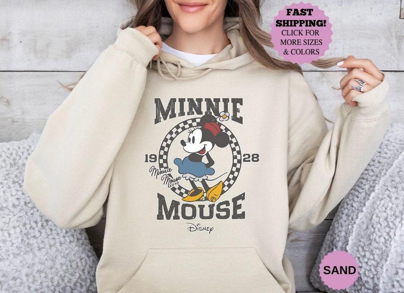 May include: Sand-colored hoodie featuring a graphic of Minnie Mouse. The design includes the text "MINNIE MOUSE" with "1928" and a checkered circle. The hoodie has a drawstring hood and a front pocket. The image also includes a pink circle with the text "FAST SHIPPING! CLICK FOR MORE SIZES & COLORS".