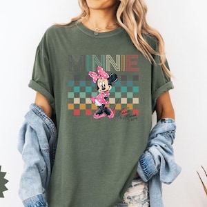 May include: Olive green t-shirt featuring a graphic of Minnie Mouse with the word "MINNIE" above her. The design includes a checkered pattern in various colors. The shirt also has the text "Minnie Mouse" in a script font.