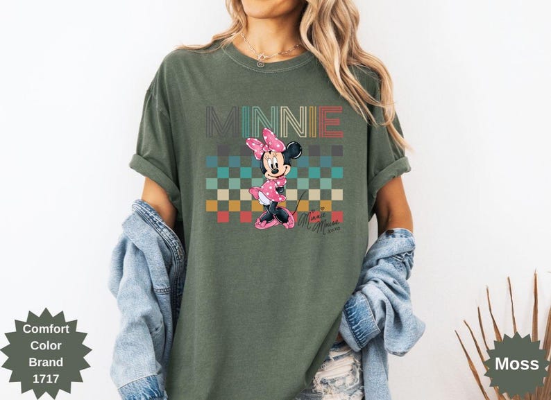 May include: Olive green t-shirt featuring a graphic of Minnie Mouse with the word "MINNIE" above her. The design includes a checkered pattern in various colors. The shirt also has the text "Minnie Mouse" in a script font.