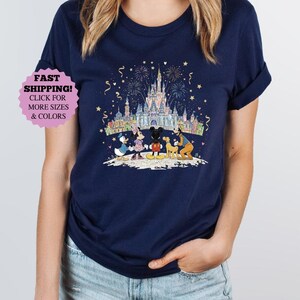 May include: Navy blue t-shirt featuring a colorful graphic of Disney characters in front of a castle with fireworks. The characters include Mickey Mouse, Minnie Mouse, Donald Duck, and Goofy. The shirt has a round neck and short sleeves.