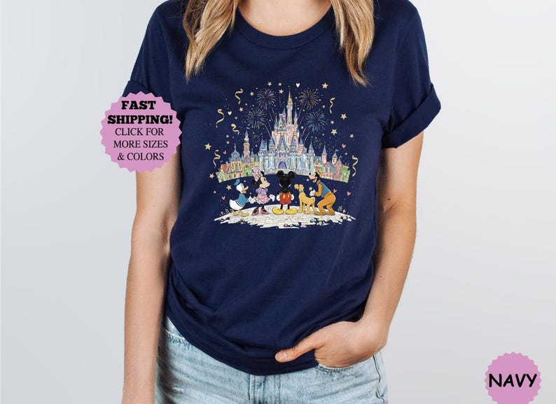 May include: Navy blue t-shirt featuring a colorful graphic of Disney characters in front of a castle with fireworks. The characters include Mickey Mouse, Minnie Mouse, Donald Duck, and Goofy. The shirt has a round neck and short sleeves.