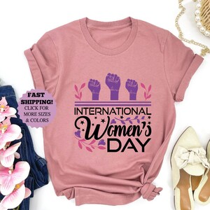 May include: Mauve-colored t-shirt with the text "International Women's Day" in black script, featuring three purple fists and floral accents. The shirt is displayed with jeans, flowers, and shoes.