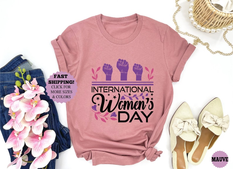 May include: Mauve-colored t-shirt with the text "International Women's Day" in black script, featuring three purple fists and floral accents. The shirt is displayed with jeans, flowers, and shoes.