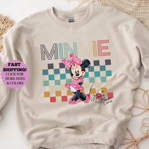 以下が含まれることがあります： ピンクのドット柄のドレスとリボンを着たミニーマウスのグラフィックが特徴のベージュのスウェットシャツ。「MINNIE」の文字が、キャラクターの上にカラフルなレトロフォントでプリントされています。ミニーの後ろには、青、緑、赤の色合いのチェック柄があります。