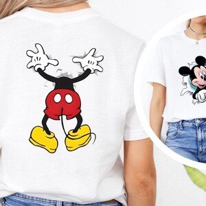 Puede incluir: Camiseta blanca con un dise&ntilde;o de Mickey Mouse. La parte delantera muestra la cara de Mickey saliendo a trav&eacute;s de un efecto rasgado, y la parte trasera muestra su cuerpo de manera similar. El dise&ntilde;o incluye los colores negro, rojo, amarillo y blanco.