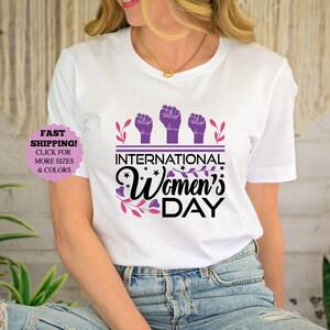 May include: White t-shirt with the text "International Women's Day" in black and pink, with purple raised fists and floral accents. The shirt is a crew neck and has short sleeves.