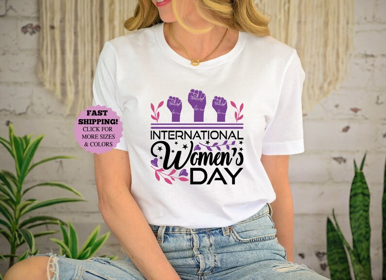 May include: White t-shirt with the text "International Women's Day" in black and pink, with purple raised fists and floral accents. The shirt is a crew neck and has short sleeves.